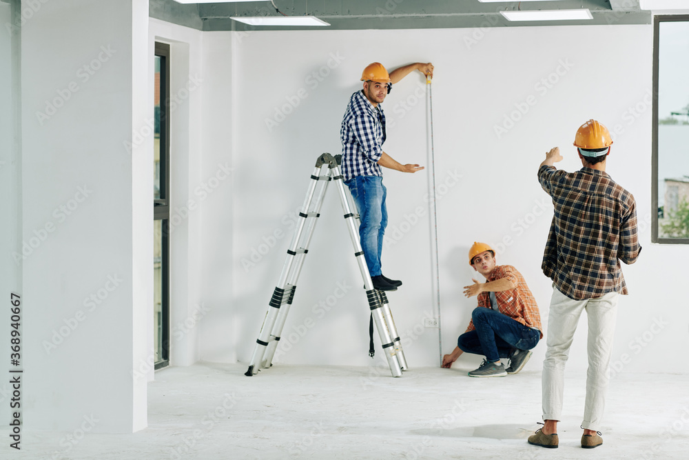 Team of construction workers in hardhats measuring wall with tape measure - obrazy, fototapety, plakaty 