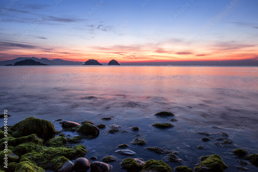 Naklejka premium Amazing seashore sunrising,endless horising and green mossy round gravel pebble beach, sea waves beats on a rocky shore with foam.