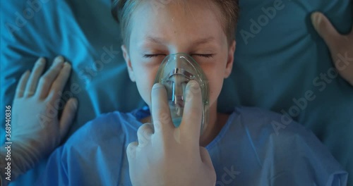 Close up of unconscious child with oxygen inhalation mask lying on medical stretcher. Paramedics checking pulse and doing cardiac massage to little boy while they going to hospital