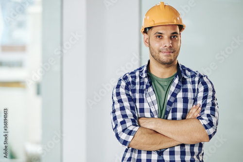 Portrait of handsome smiling contractor in hardhat folding arms and looking at camera