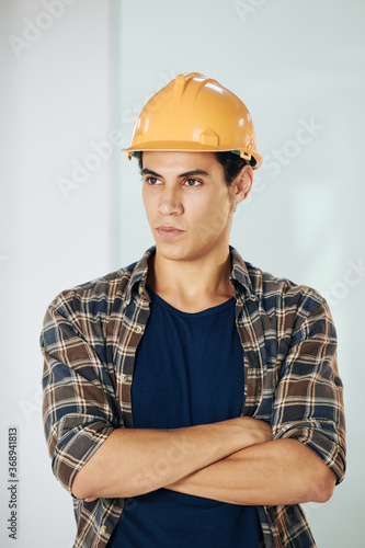 Portrait of handsome serious young mixed-race young builder in hardhat crossing arms and looking away