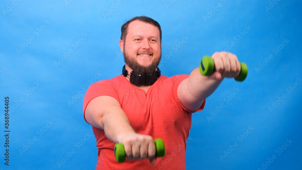 fat fancy man in a pink t-shirt with dumbbells funny doing exercises on a blue background.