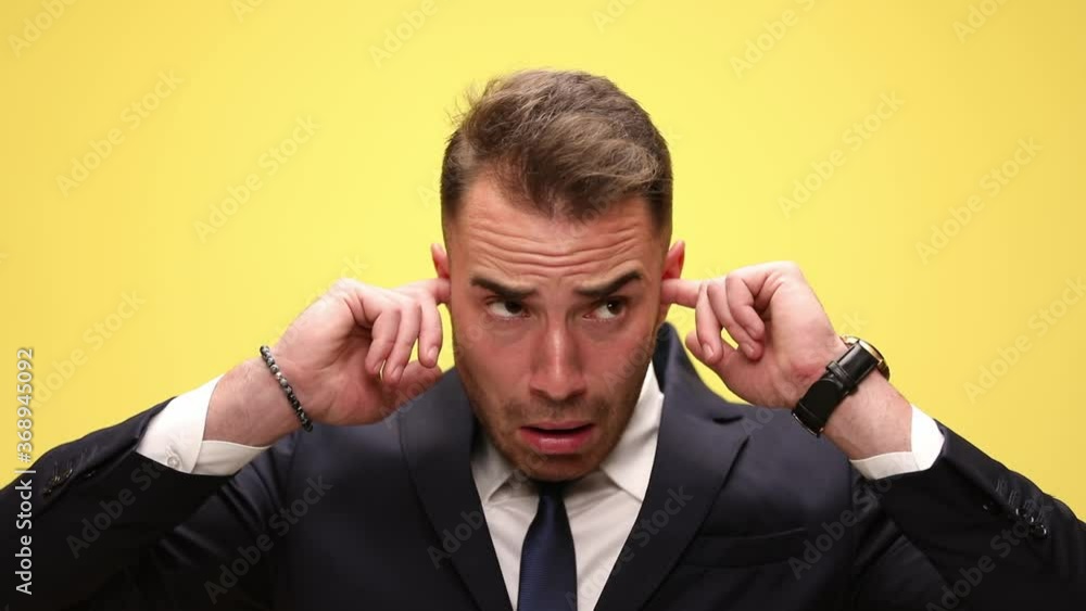 elegant young businessman in suit covering ears with fingers stopping the noise, making funny faces and smiling on yellow background elegant young businessman in suit covering ears with fingers stopping the noise, making funny faces and smiling on yellow background