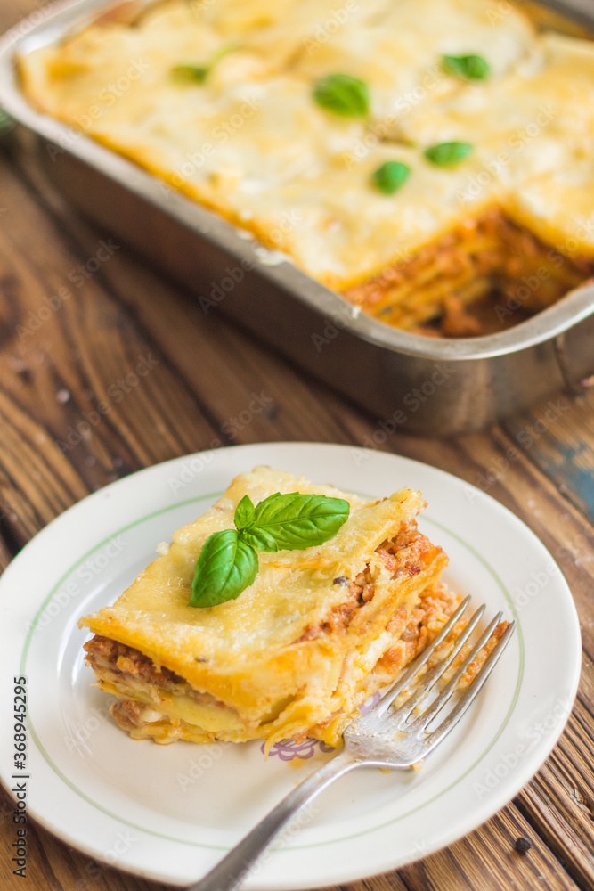 Homemade lasagna ready to eat in metal baking tray served with basil leaves, tomatoes, and turquoise tablecloth on brown wooden background. One portion on the white plate with fork. Top view. Homemade lasagna ready to eat in metal baking tray served with basil leaves, tomatoes, and turquoise tablecloth on brown wooden background. One portion on the white plate with fork. Top view.
