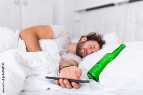 Texting while drunk concept. Young man lying in bed deadly drunken holding empty bottle of booze. intoxicated with alcohol . Alcoholism habitual drunkenness pernicious habit concept