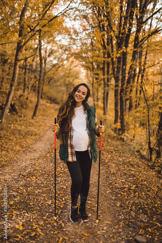 Young happy pregnant woman hiking in autumn forest with trekking poles. Nordic walking and fitness. Pregnancy activity and healthy lifestyle concept.