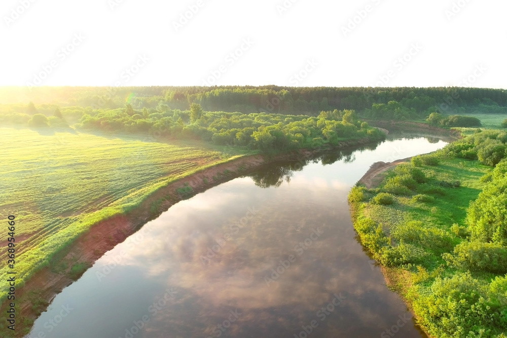 Flying over river and green forest at summer sunny day, sky reflecting in water. Sunrise on nature in beautiful place with natural landscape. Aerial view of picturesque woodland and river.