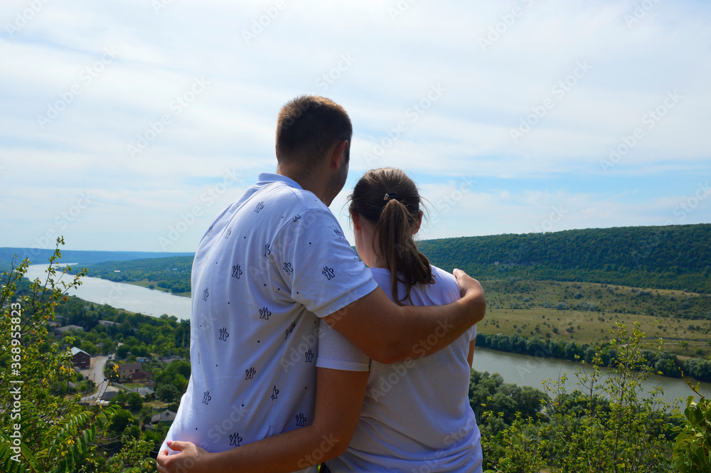 a couple in love stands hugging on the hillside and look down at the river, forest and a small village