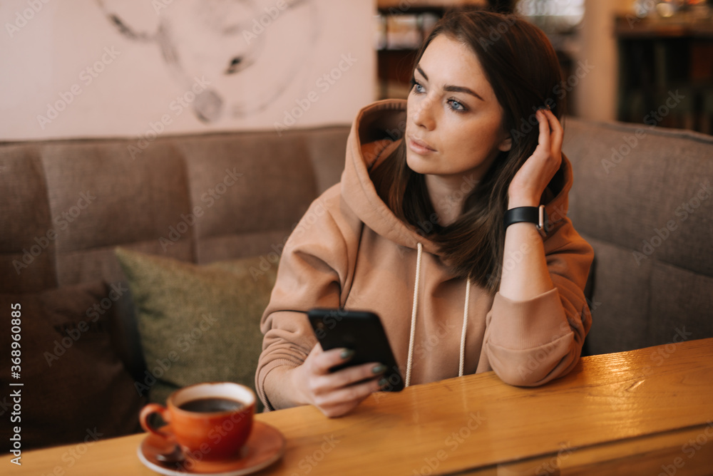 Portrait of young attractive woman typing message in phone at cozy coffee shop. Pretty girl in casual clothes thinks while typing on the phone at cafe. Concept of leisure activity. Portrait of young attractive woman typing message in phone at cozy coffee shop. Pretty girl in casual clothes thinks while typing on the phone at cafe. Concept of leisure activity.
