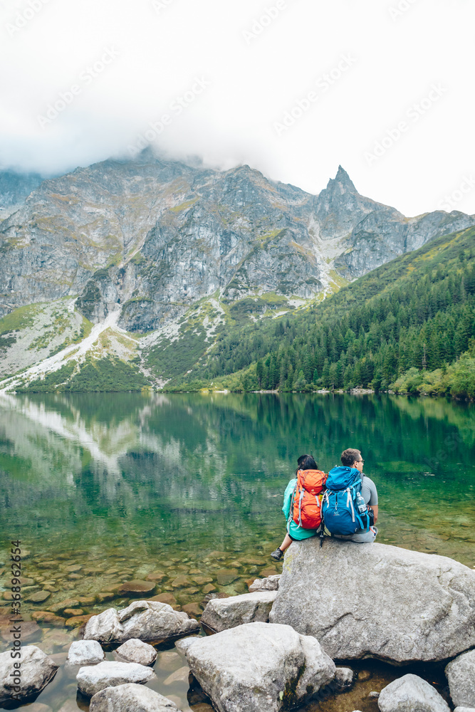 Morske Oko, Poland - September 12, 2019: hikers couple sitting on rock looking at lake in mountains Morske Oko, Poland - September 12, 2019: hikers couple sitting on rock looking at lake in mountains