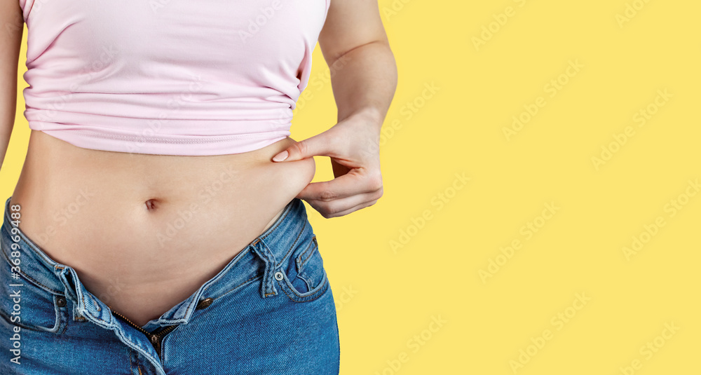 Woman in unbuttoned jeans holds with hand and squeezes excess belly fat.  The concept of overweight, weight loss, diet, obesity, junk food on yellow  background, banner, copy space. Photos | Adobe Stock
