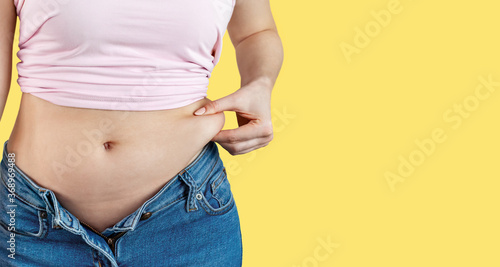 Woman in unbuttoned jeans holds with hand and squeezes excess belly fat. The concept of overweight, weight loss, diet, obesity, junk food on yellow background, banner, copy space. © KseniyA