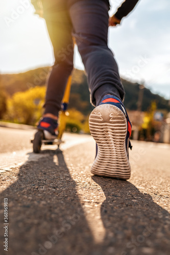 A teenage boy rides a scooter. Right foot in a sneaker close-up. Bottom and rear view. Empty street in Sunny weather in the background. Concept of youth activity  sports and recreation