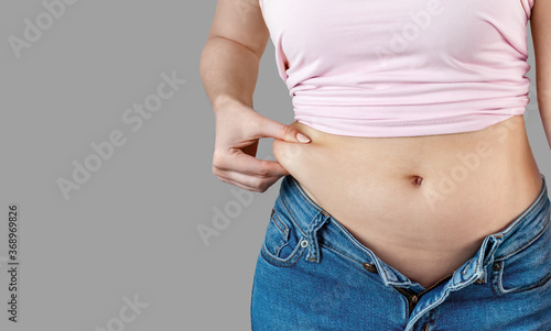 Woman in unbuttoned jeans holds with hand and squeezes excess belly fat. The concept of overweight, weight loss, diet, unhealthy lifestyle, obesity, junk food on gray background, banner, copy space. © KseniyA