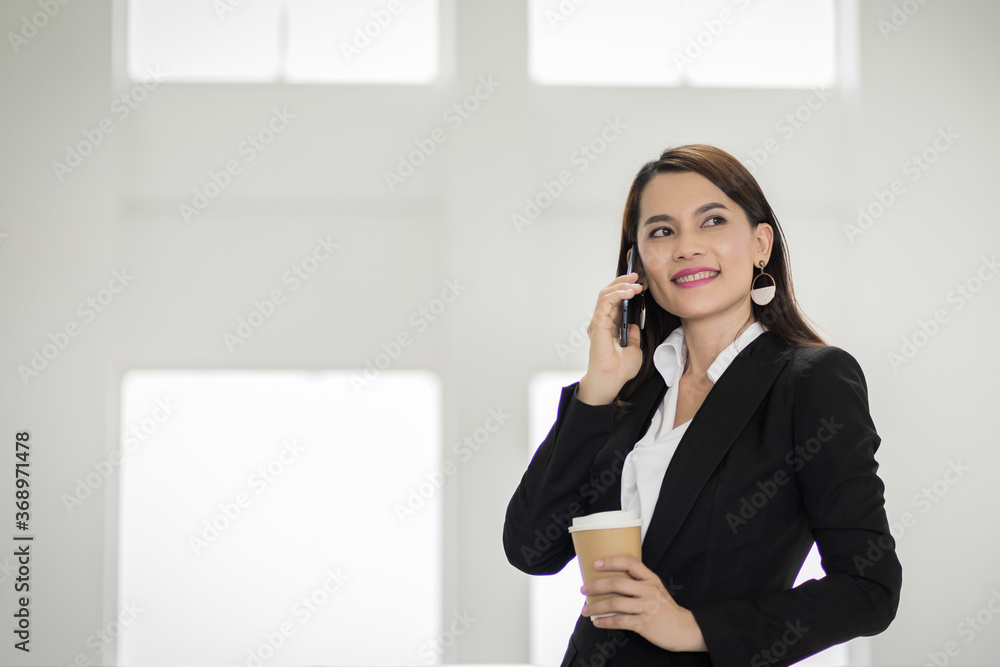 Portrait of a young Asian business woman talking over cellphone and holding cup of coffee in business building. Photo of beautiful girl in casual suite with phone and cup of coffee. stock photo Portrait of a young Asian business woman talking over cellphone and holding cup of coffee in business building. Photo of beautiful girl in casual suite with phone and cup of coffee. stock photo