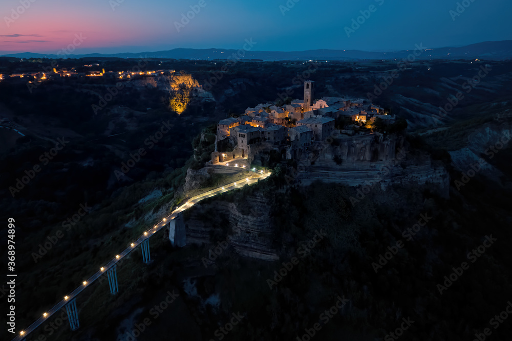 Fototapeta premium Civita di Bagnoregio. Aerial, night view of ancient Italian city standing on top of a plateau, illuminated by lamps. Famously known as dying city. City on steep rock over Tiber river valley, Italy.