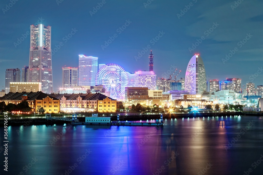 Vibrant night scenery of Yokohama Minatomirai Bay Area with Landmark Tower among skyscrapers in background, a giant Ferris wheel in an amusement park & reflections of colorful city lights on water