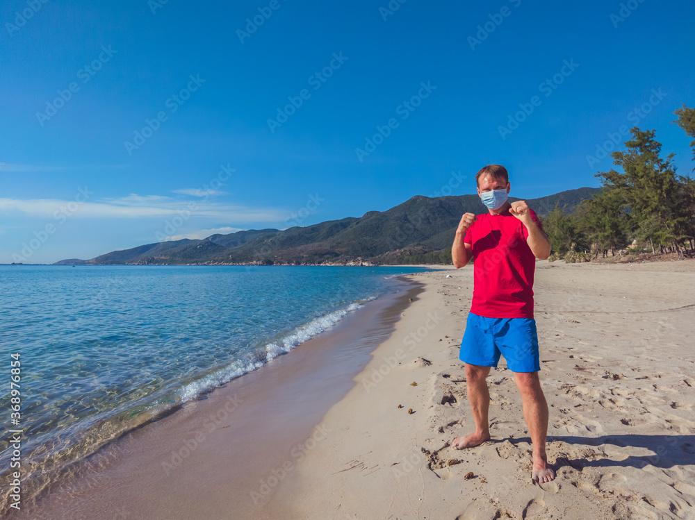 Young muscle man in medical mask perform outdoor workouts in the park near sea during second wave coronavirus Covid 19 pandemic quarantine. Kick training pose, swing arms and legs, sport active life