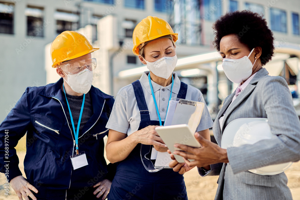 Black engineer and two workers with face masks cooperating while using digital tablet at construction site. Black engineer and two workers with face masks cooperating while using digital tablet at construction site.