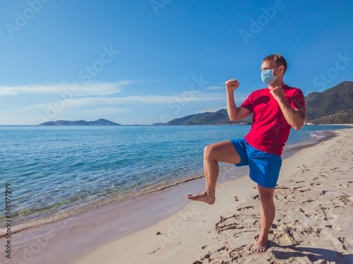 Young muscle man in medical mask perform outdoor workouts in the park near sea during second wave coronavirus Covid 19 pandemic quarantine. Kick training pose, swing arms and legs, sport active life