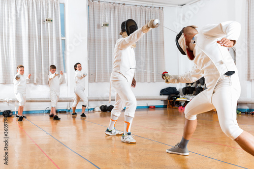 Positive smiling coaches demonstrating to young athletes attack movements with rapier during fencing workout