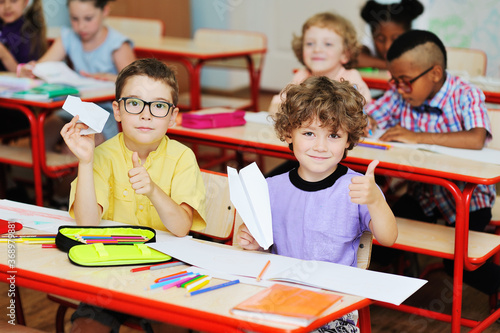 two schoolboy boys with paper planes in their hands smile against the background of a group of children in a school classroom. Back to school, September 1, knowledge day