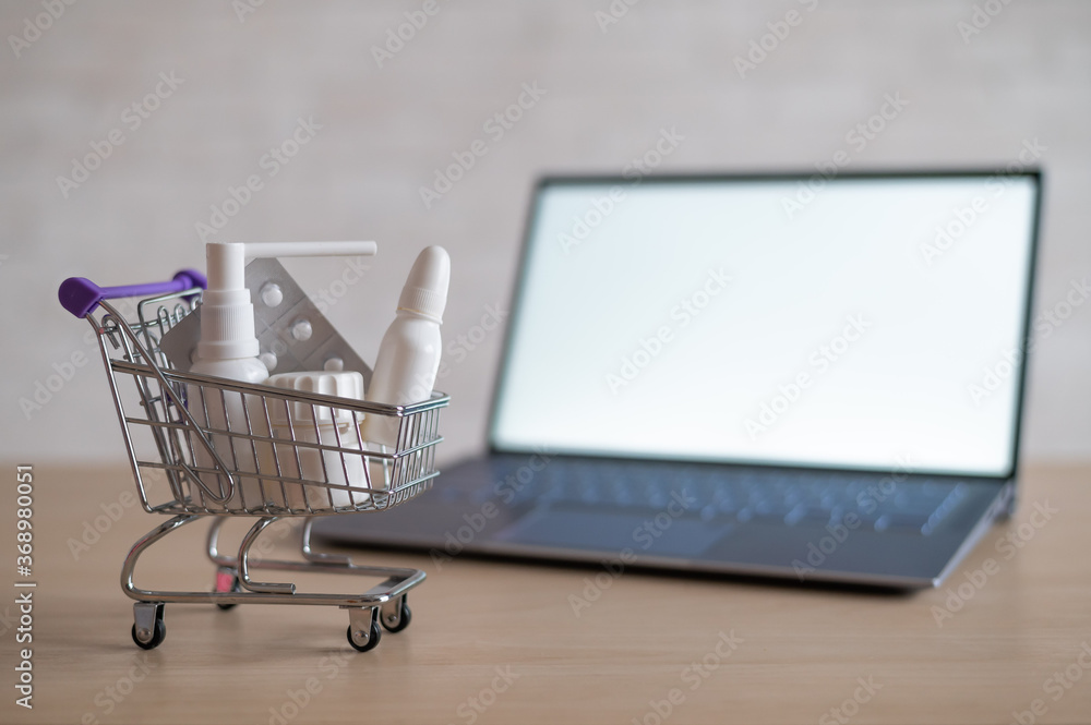 Laptop with blank white screen and miniature shopping cart full of medicines. Online pharmacy concept. Website for buying tablets.