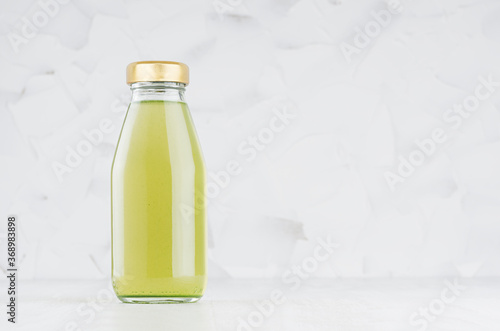 Green kiwi juice in glass bottle with gold cap mock up on white wood table in light interior with copy space, template for packaging, advertising, design product, branding.