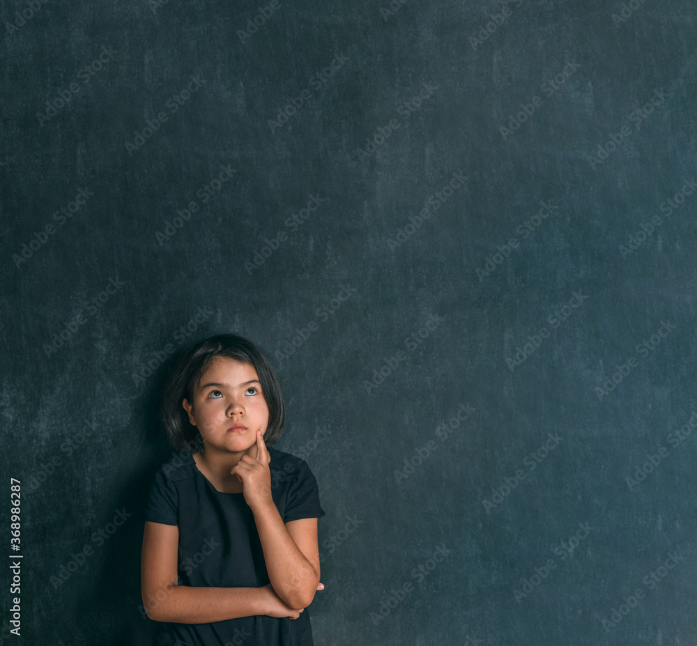 Back to school. Serious little girl thinking touching her head with a finger near blackboard. Child from elementary school. Education. Back to school. Serious little girl thinking touching her head with a finger near blackboard. Child from elementary school. Education.