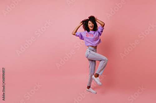 Full-length shot of attractive stylish woman dancing on rosy background. Appealing female model in jeans fooling around in studio.