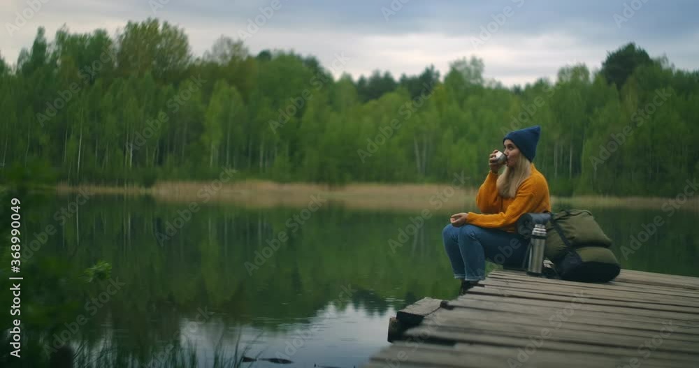A female traveler drinks tea with a backpack and enjoys the beauty of a mountain forest lake sitting on a wooden pier. Slow-motion concept of a lonely woman's solitary journey. Quarantine and travel A female traveler drinks tea with a backpack and enjoys the beauty of a mountain forest lake sitting on a wooden pier. Slow-motion concept of a lonely woman's solitary journey. Quarantine and travel