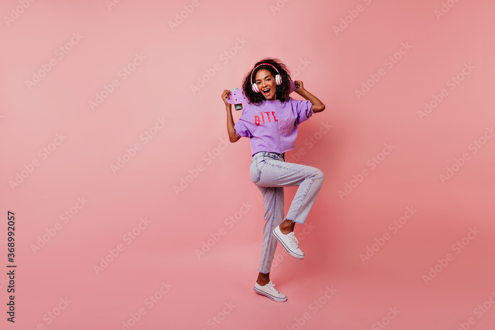Lovely girl in white shoes dancing in studio while listening music. Full-length photo of refined african lady with skateboard chilling on pink background.