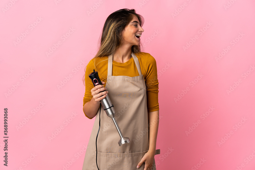 Young chef woman using hand blender isolated on pink background laughing in lateral position