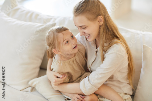Warm toned high angle portrait of smiling teenage girl holding cute little sister while sitting on cozy white couch at home, copy space