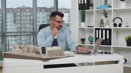 Good-looking balanced experienced bearded architect in glasses checking technical measurements from computer on wooden model of new building in design office ,4k photo