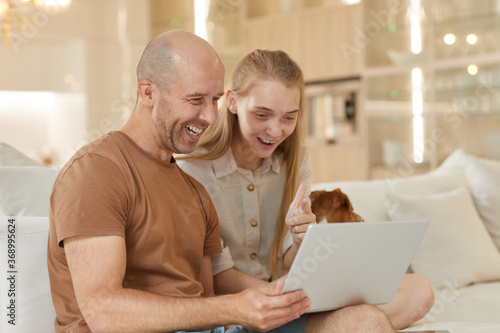 Warm-toned portrait of mature father smiling happily while looking at laptop screen with teenage girl while sitting on couch in home interior, copy space