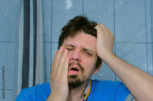 Depressed shaggy young man with headache and hangover in bathroom in morning rubs his face. Portrait of sick guy with flu, cold looking at camera as at mirror. Midlife crisis, depression and stress. photo