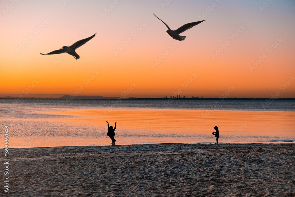 Silhouettes of children playing on ocean beach at sunset with birds flying and city skyscrapers in the distance