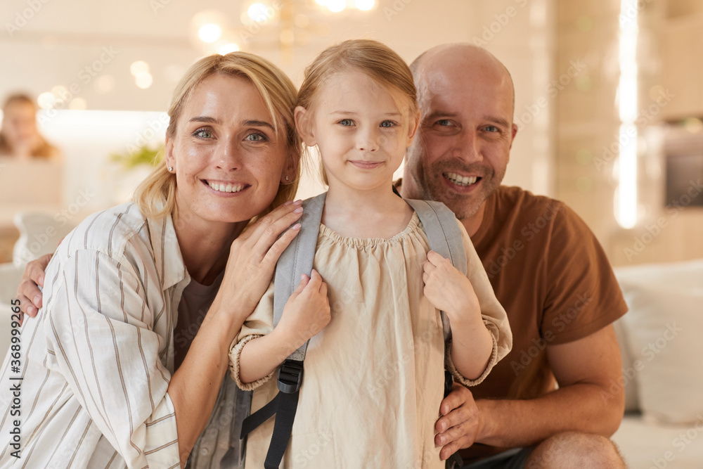 Warm-toned portrait of happy family looking at camera and smiling while posing with cute little girl carrying backpack Warm-toned portrait of happy family looking at camera and smiling while posing with cute little girl carrying backpack