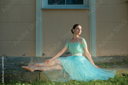 Beautiful graceful young ballerina in pointe shoes and blue dress sitting on the pavement. Female stretching indoors, professional arts. Female fit and healthy lifestyle. Ballet practice on sunset