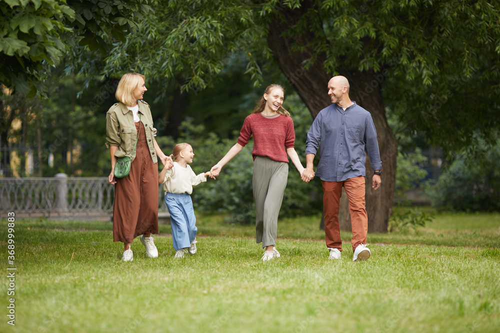 Full length portrait of modern carefree family with two kids holding hands while walking towards camera on green grass outdoors, copy space