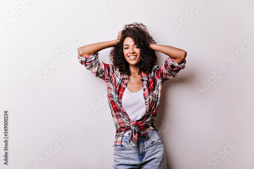 Shapely slim lady in casual attire touching her wavy hair. Appealing african girl standing on white background.