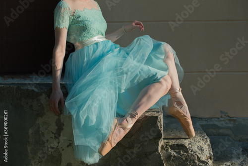 Beautiful graceful young ballerina in pointe shoes and blue dress sitting on the stairst. No face female feet legs stretching indoors toes professional arts. Ballet practice and stretching