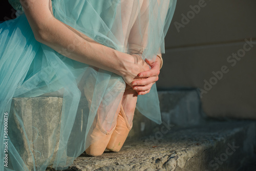 Faceless ballet dancer in pointe shoes and blue dress sitting on the stairs. Dancing female feet legs stretching outdoors professional arts. Hands and legs of ballet dancer. Close-up view, copy space