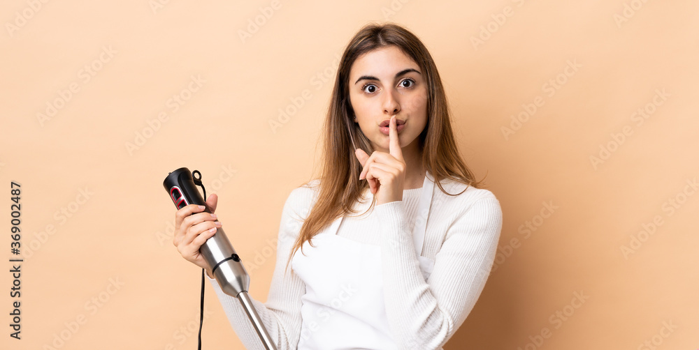 Woman using hand blender over isolated background showing a sign of silence gesture putting finger in mouth Woman using hand blender over isolated background showing a sign of silence gesture putting finger in mouth
