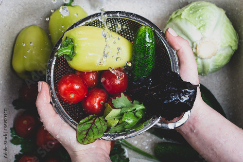 women's hands hold a metal colander full of fresh ripe vegetables and herbs under a spray of water over the kitchen sink where the rest of the vegetables are stored