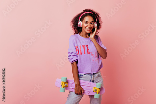 Amazing african woman in purple sweater posing with skateboard. Debonair black female model standing on pink background in headphones.