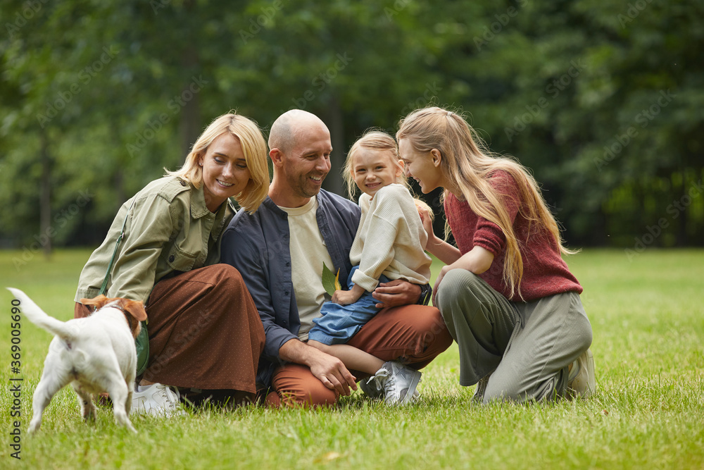 Full length portrait of happy modern happy family with two daughters and pet dog sitting on green grass outdoors and enjoying time in park together, copy space