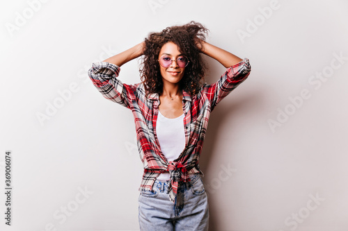 Winsome african woman in glasses playing with her curly hair. Indoor photo of debonair black girl in stylish checkered shirt.