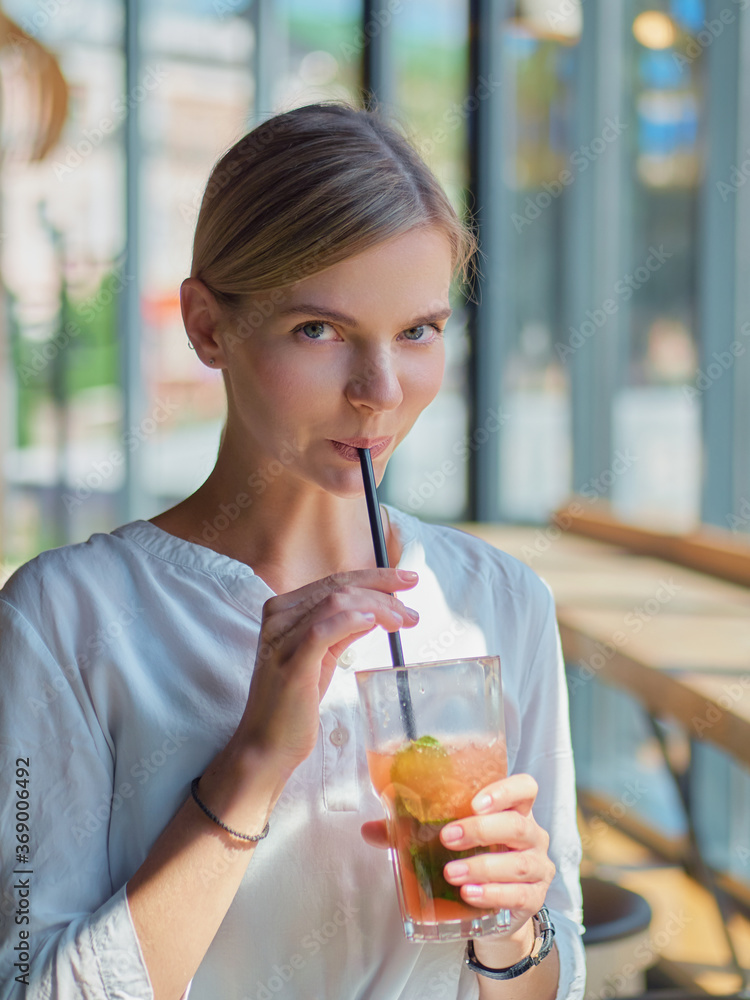 Calm beautiful happy young woman in relaxed mood looking outdoors and smiling sitting at the bar counter near big window with healthy delicious fresh lemonade glass dressed trendy white shirt Calm beautiful happy young woman in relaxed mood looking outdoors and smiling sitting at the bar counter near big window with healthy delicious fresh lemonade glass dressed trendy white shirt